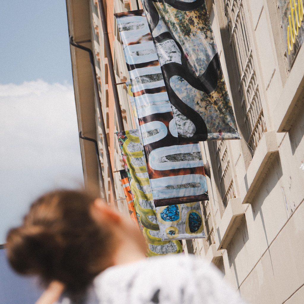 Progetto Playing Ground: donna in primo piano guarda le bandiere illustrate sulla facciata del centro culturale poli-funzionale BASE Milano.